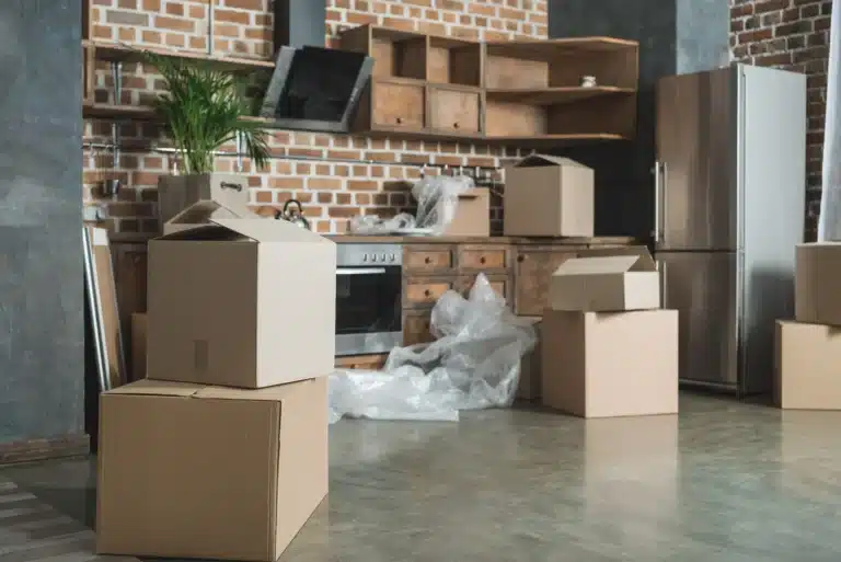 Cardboard boxes stacked in a partially unpacked kitchen, indicating a recent move. The room features exposed brick, wooden cabinets, and stainless steel appliances. Bubble wrap and packing materials are scattered on the floor, suggesting ongoing relocation efforts by local movers.