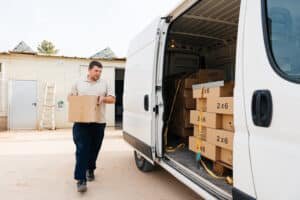 A delivery driver, wearing a beige polo shirt and dark pants, carefully carries a cardboard box from a white cargo van. The van is loaded with additional boxes marked "2x6," suggesting a local moving or delivery service.
